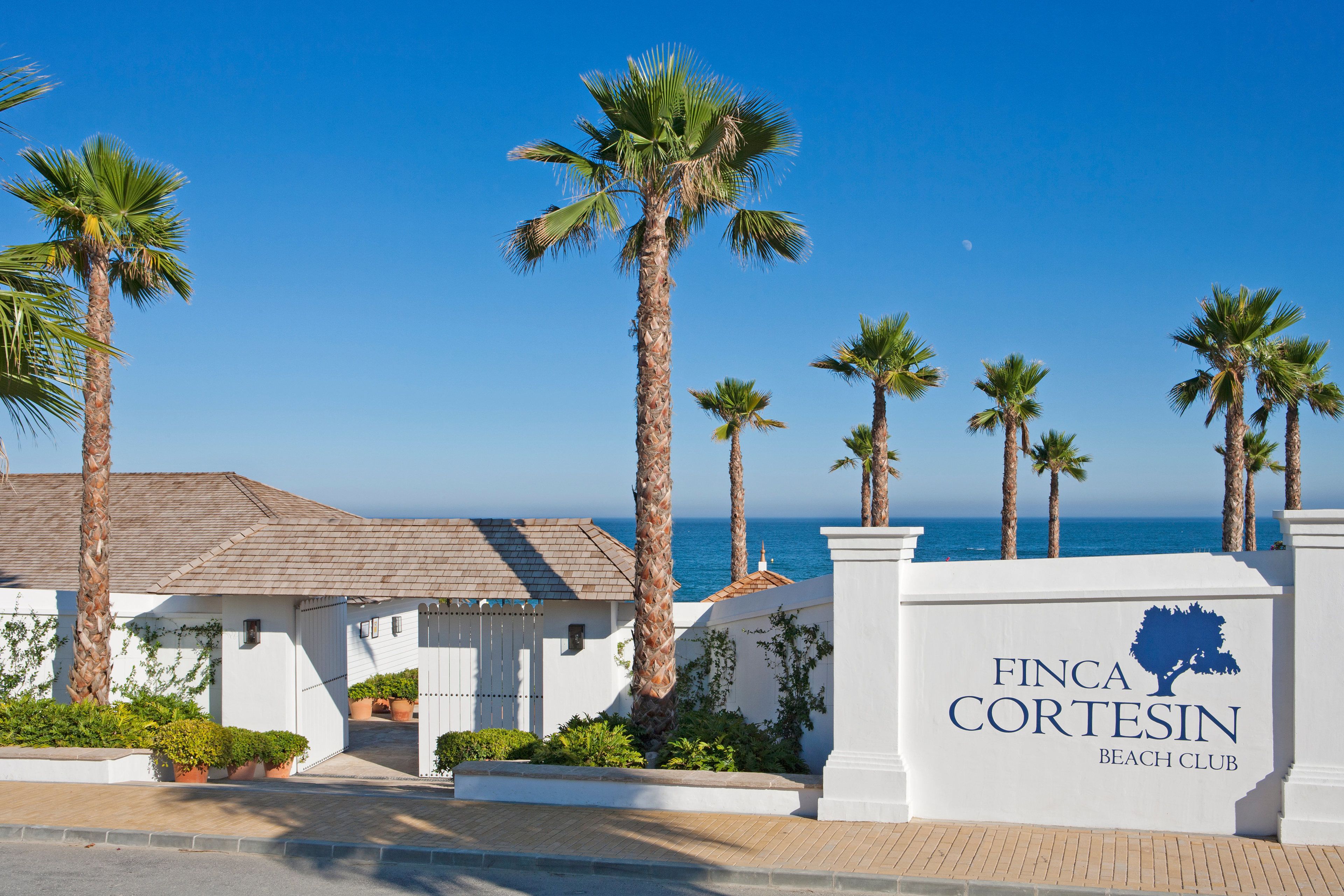 Propiedad frente a la playa con palmeras, cielo despejado y un cartel para Finca Cortesín Beach Club.