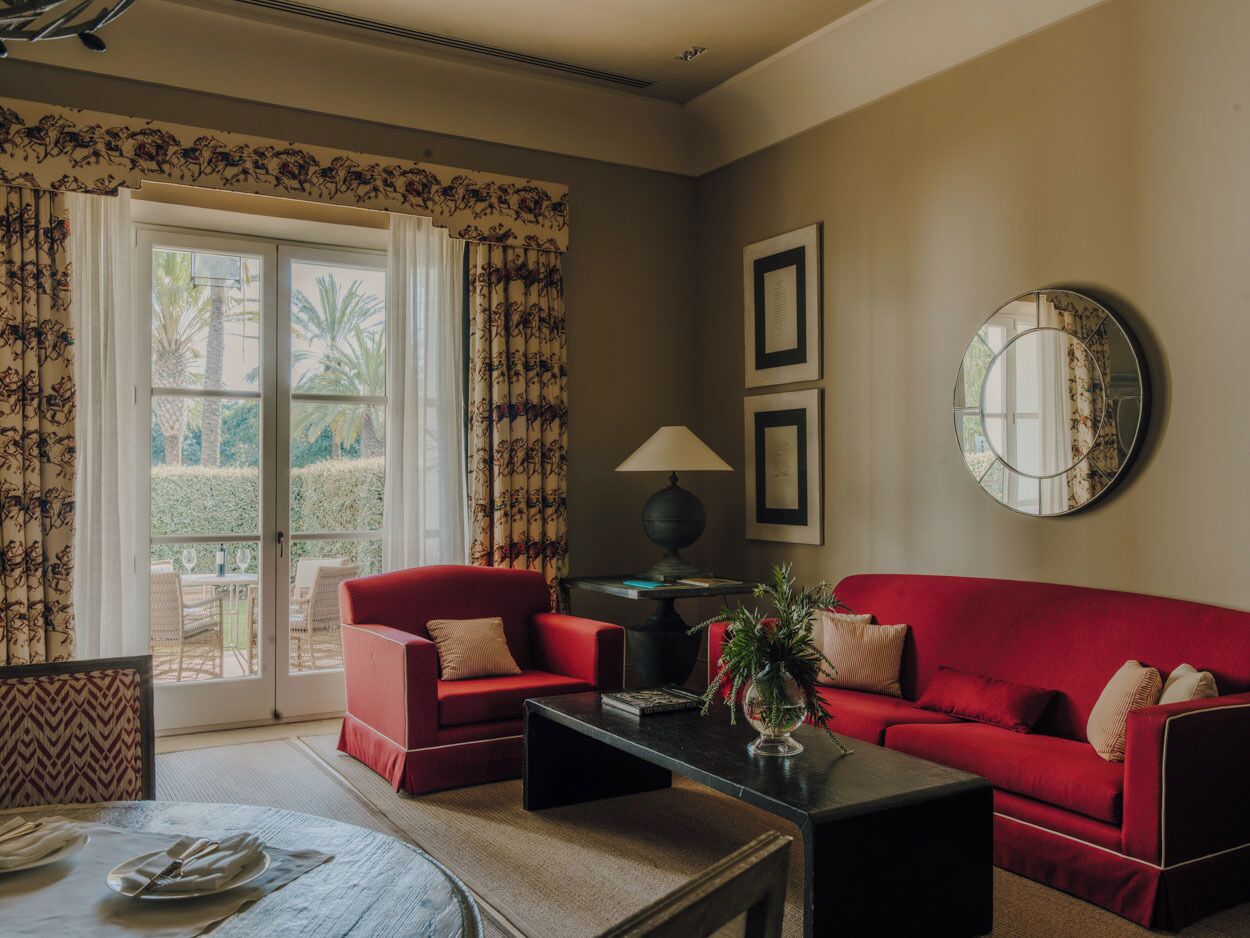 Un salon avec un canapé rouge, une table basse et une lampe. Il y a une fenêtre avec des rideaux, et de l’extérieur, on peut voir de la verdure.