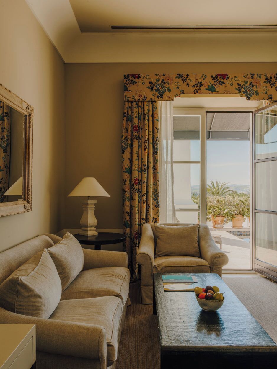 A room with a sofa, armchair, and a coffee table with a fruit bowl. There is a window with curtains and a view of the outdoors.