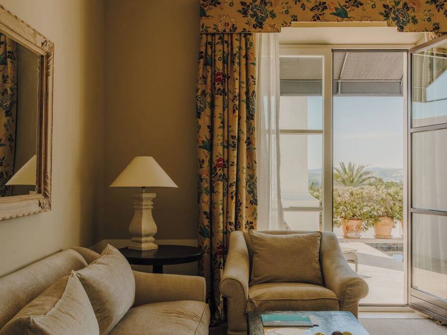 A room with a sofa, armchair, and a coffee table with a fruit bowl. There is a window with curtains and a view of the outdoors.