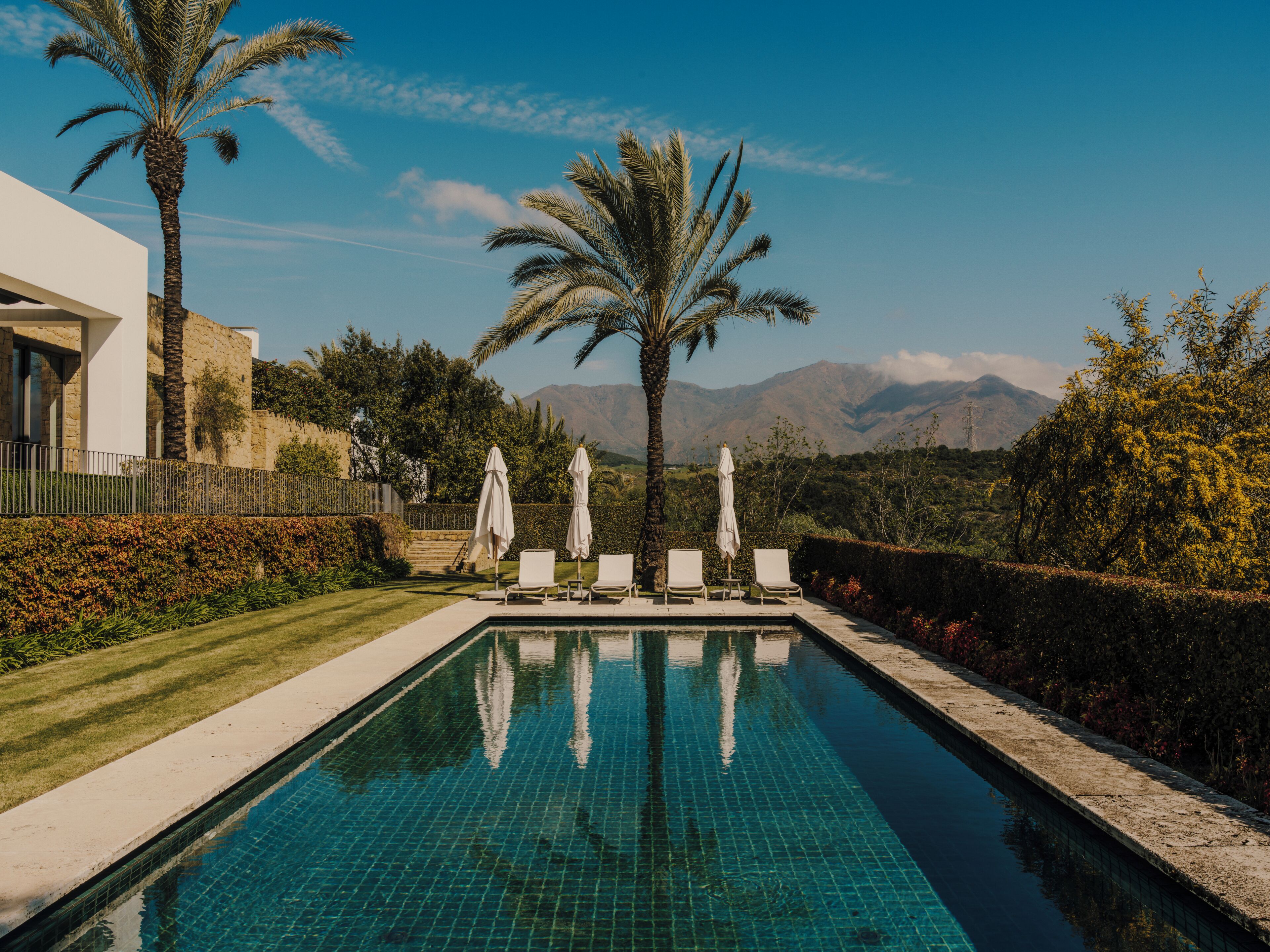 A swimming pool with lounge chairs and umbrellas, surrounded by palm trees and a view of mountains.