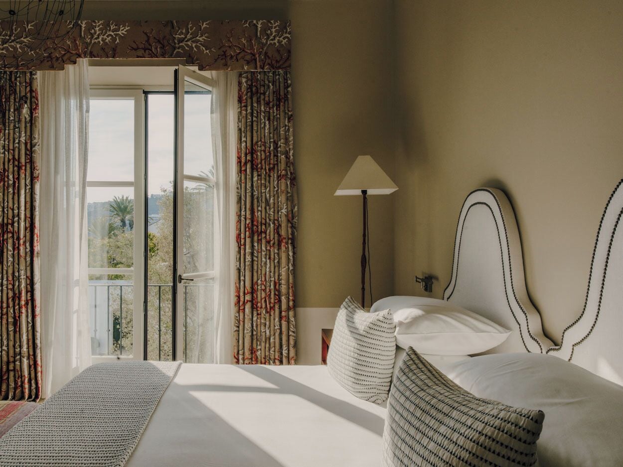 Habitación de hotel con cama, mesita de noche, lámpara y vista a través de una ventana con cortinas.