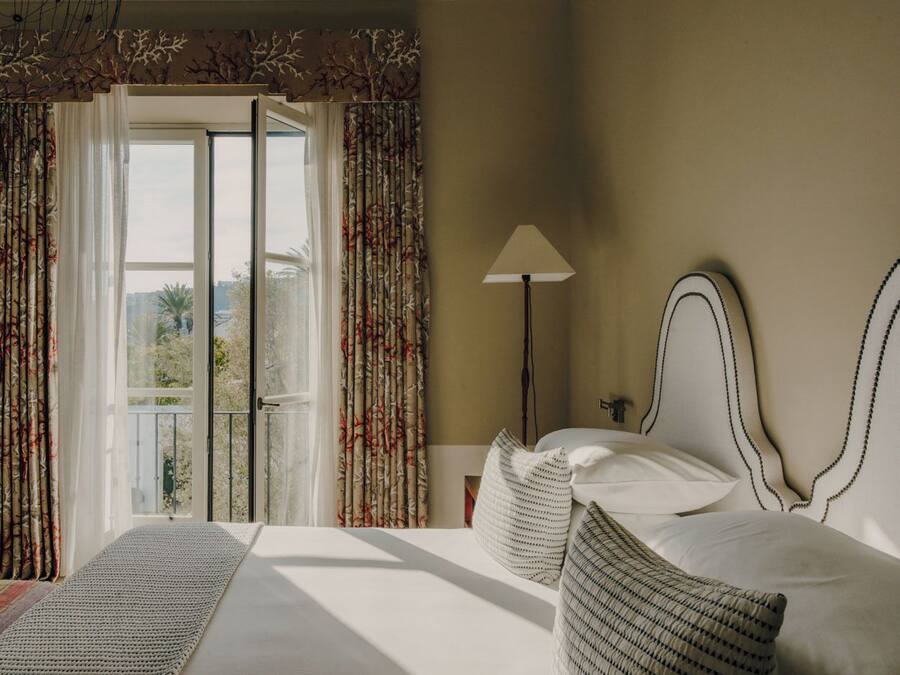 Habitación de hotel con cama, mesita de noche, lámpara y vista a través de una ventana con cortinas.