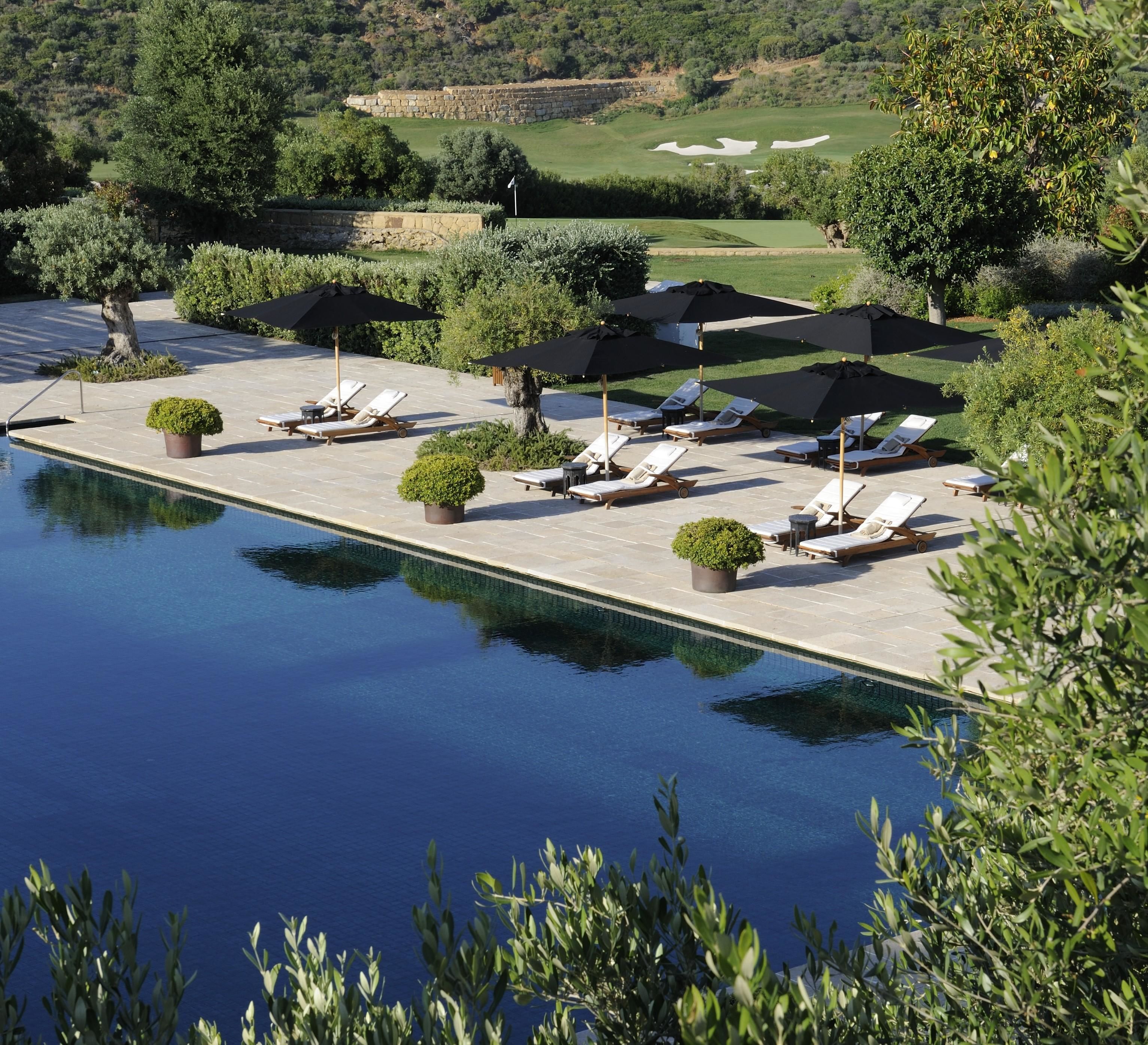 Zona de piscina con sillones de descanso y sombrillas, rodeada de vegetación y vistas a un campo de golf.