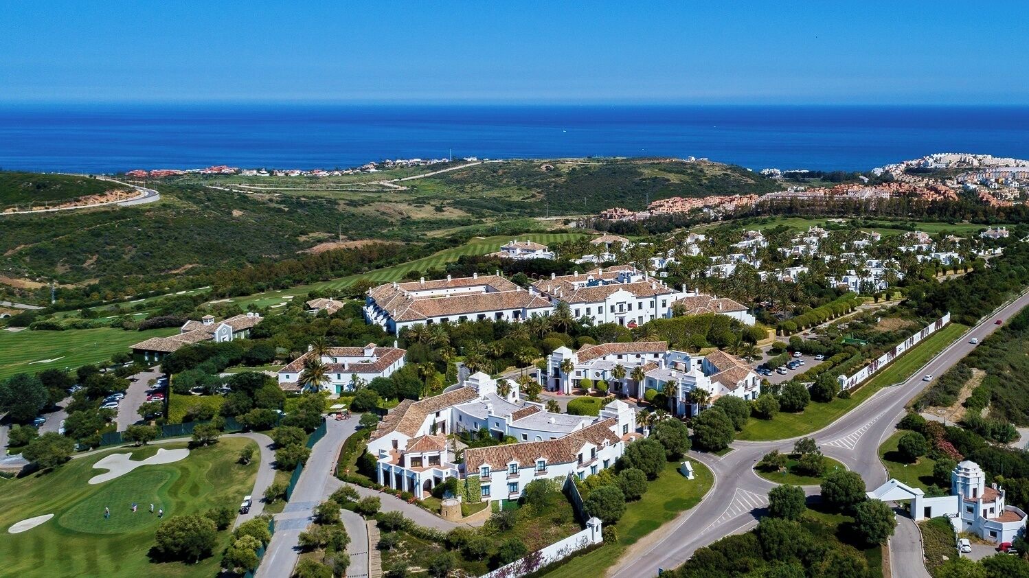 Vue aérienne d’un quartier résidentiel situé sur la côte, avec des bâtiments blancs, des pelouses verdoyantes et une route qui serpente à travers le paysage.