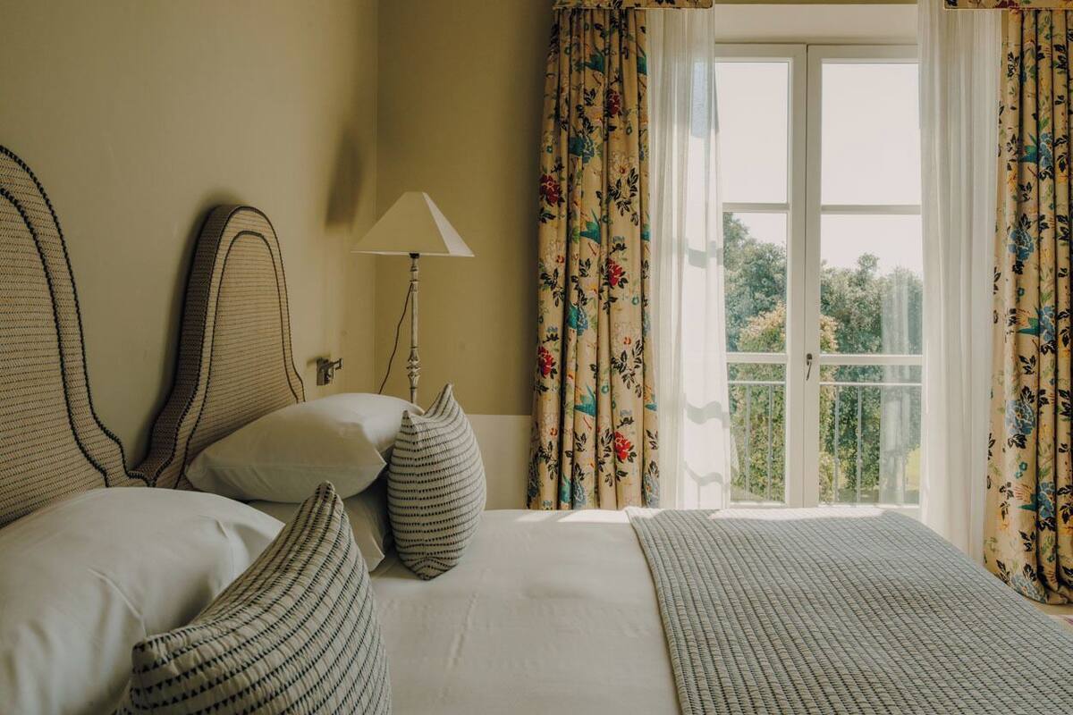 Habitación de hotel con una cama grande, dos almohadas, una lámpara de noche y un ventanal con cortinas florales.