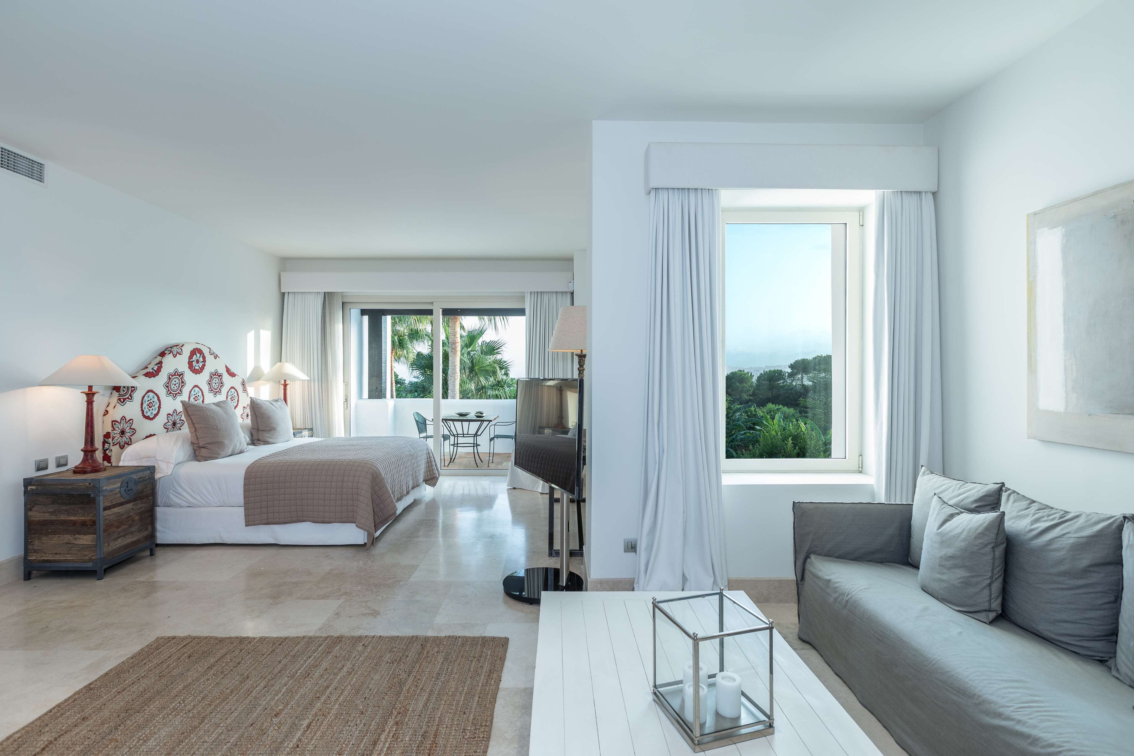 A modern hotel room with a bed, a sofa, a TV, and a balcony with a view of palm trees.
