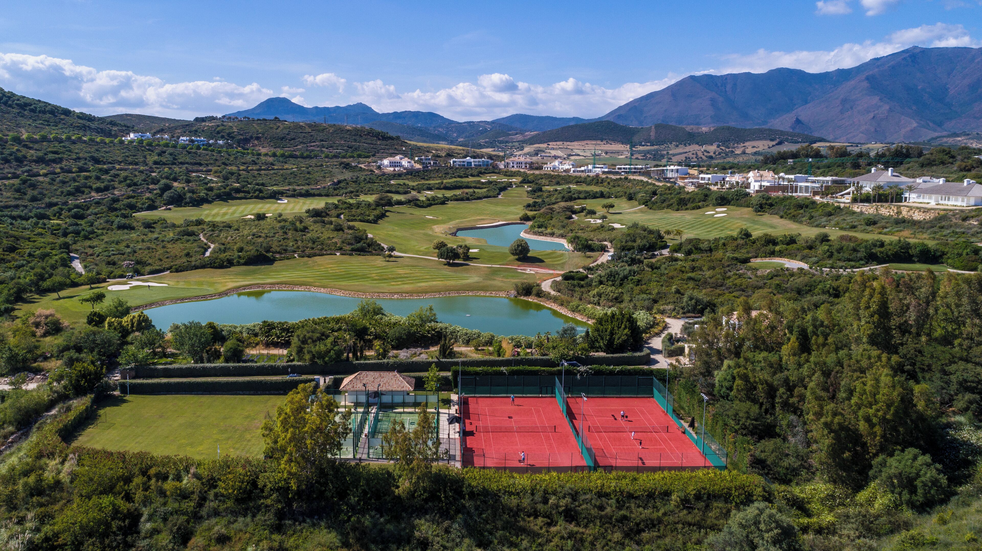 Campo de golf con varios estanques y una cancha de tenis, rodeado de abundante vegetación y montañas al fondo.