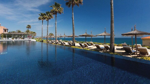 A poolside area with lounge chairs and palm trees, overlooking a clear blue sea.