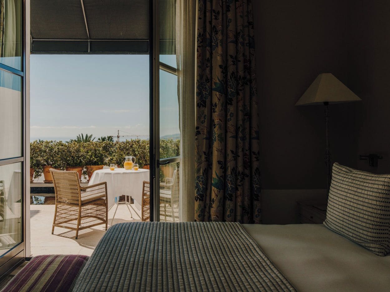 Una camera d'albergo con un letto, un tavolo con sedie, vista all'esterno e una finestra con tende.