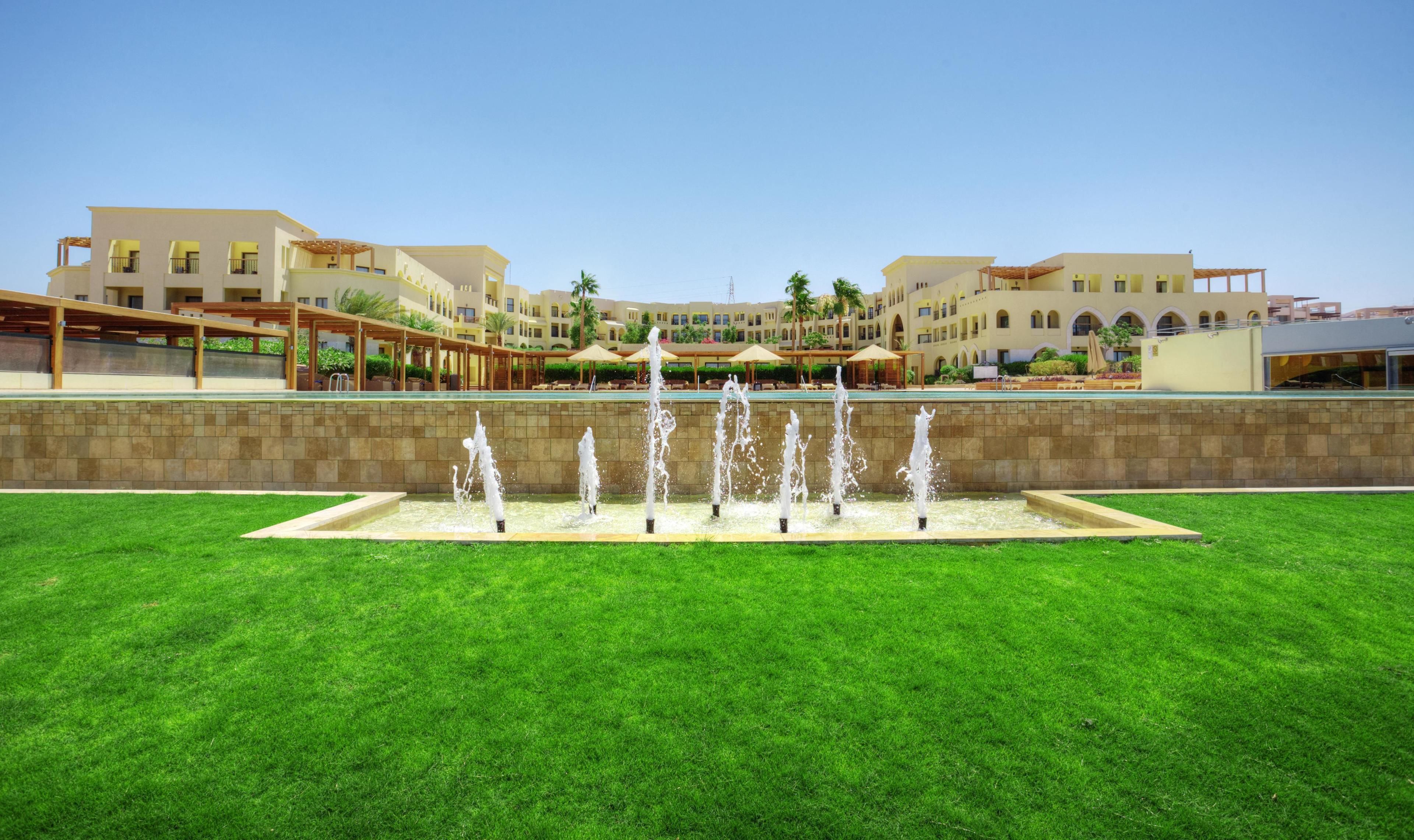 A hotel courtyard with a central fountain, surrounded by a well-maintained lawn and a pool area.