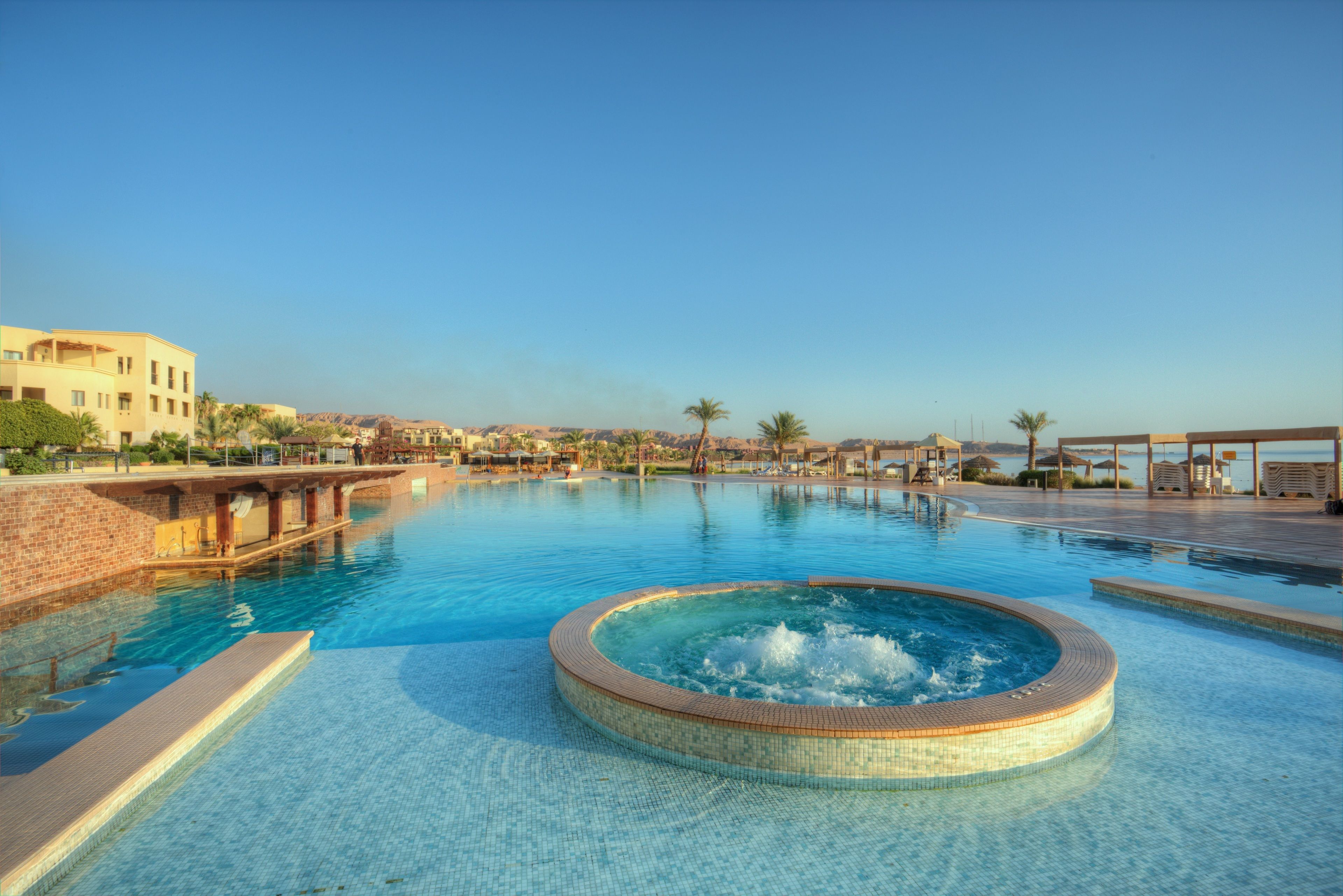 Una gran piscina al aire libre con un jacuzzi en el centro, rodeada de zonas de descanso y palmeras.
