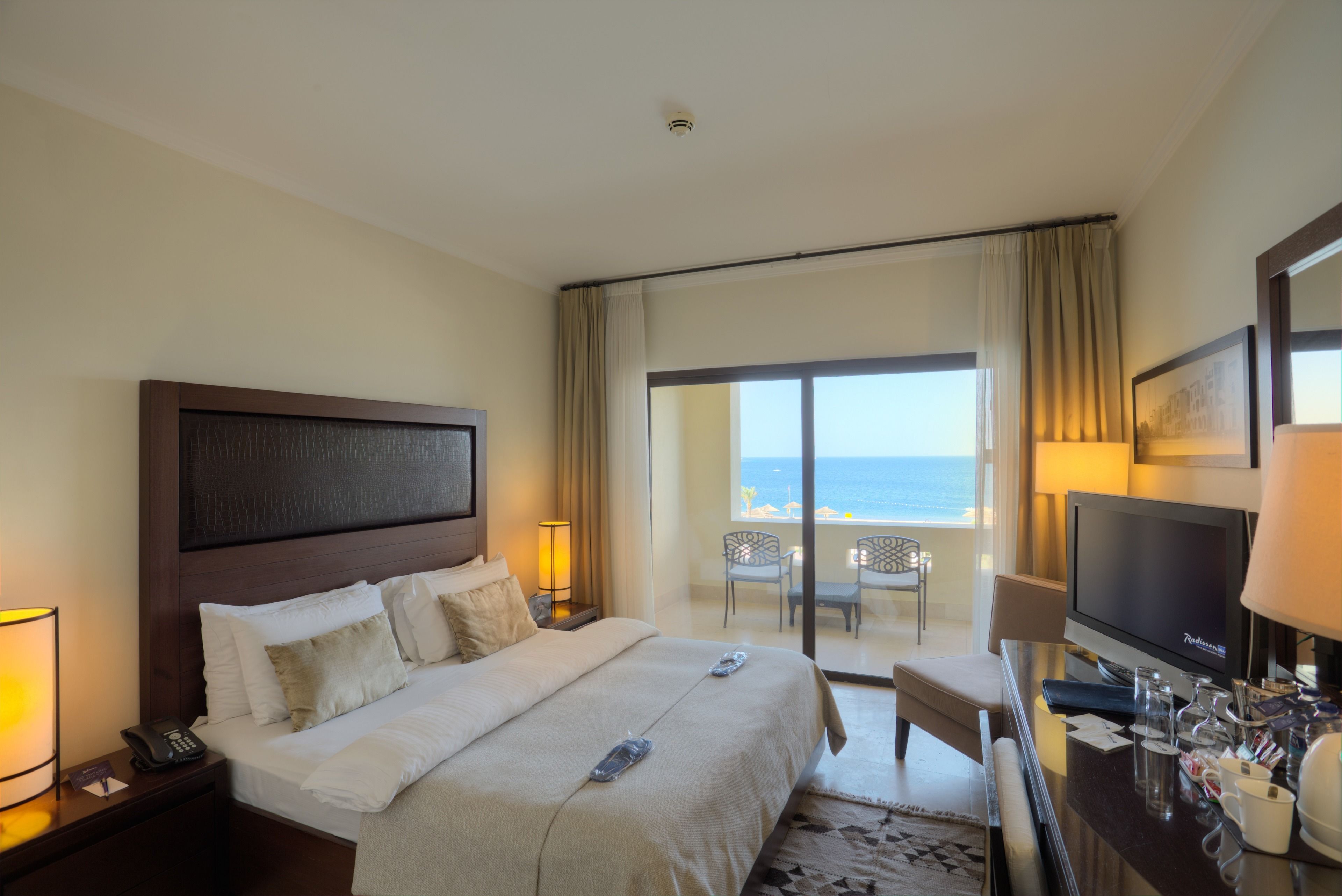 Habitación de hotel con una cama grande, televisor, escritorio con lámpara y balcón con vista al océano.