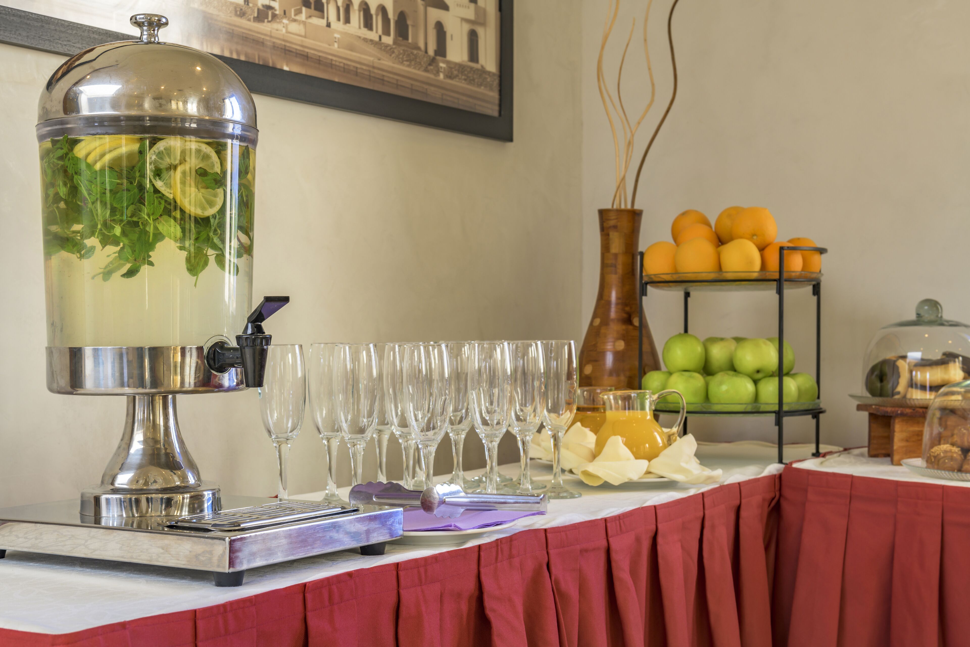 Disposición de buffet con dispensador de bebidas, exhibición de frutas y un soporte para copas de vino.