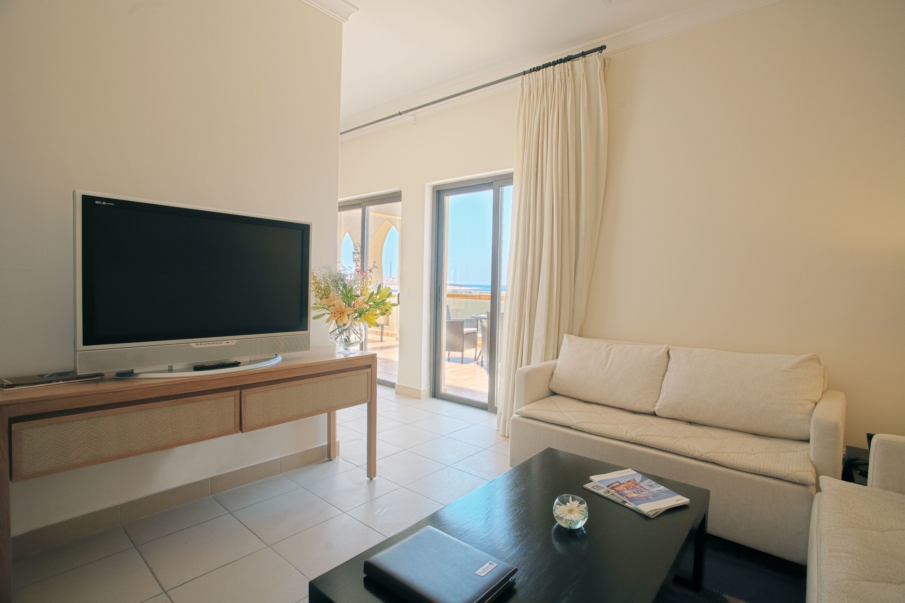 A modern living room with a flat-screen TV, a sofa, a coffee table, and a view of the outdoors through a sliding glass door.