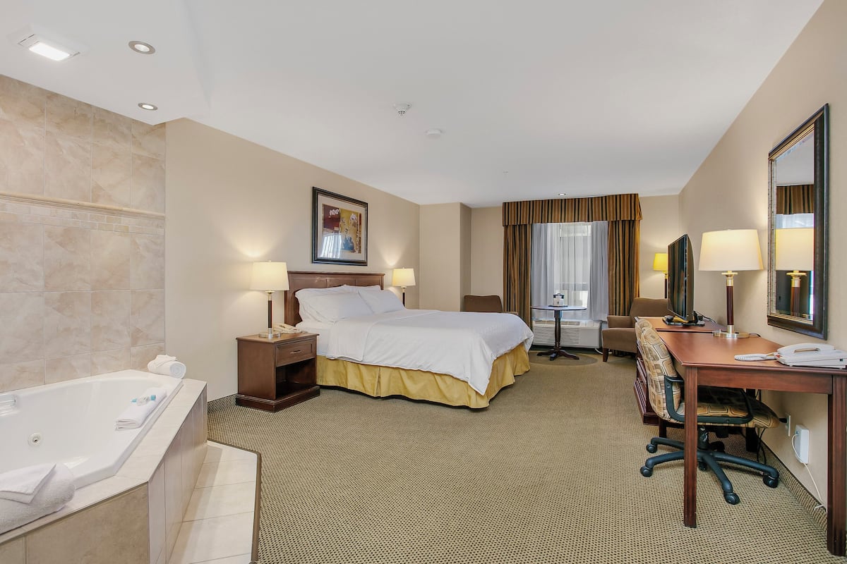 A hotel room with a large bed, a desk, a chair, a desk lamp, a TV, a window with curtains, a bathroom with a bathtub, and a mirror.