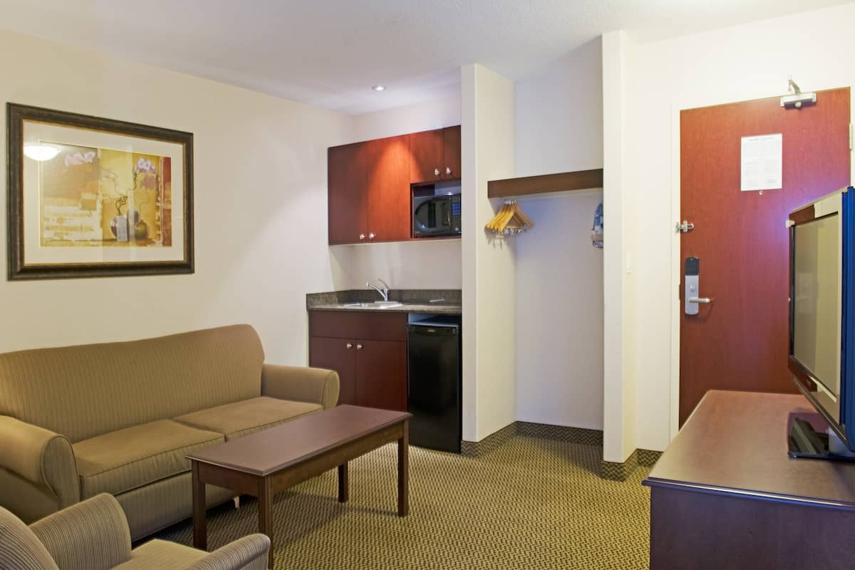 A compact hotel room with a sofa, a small dining table, a kitchenette with a microwave, and a television.