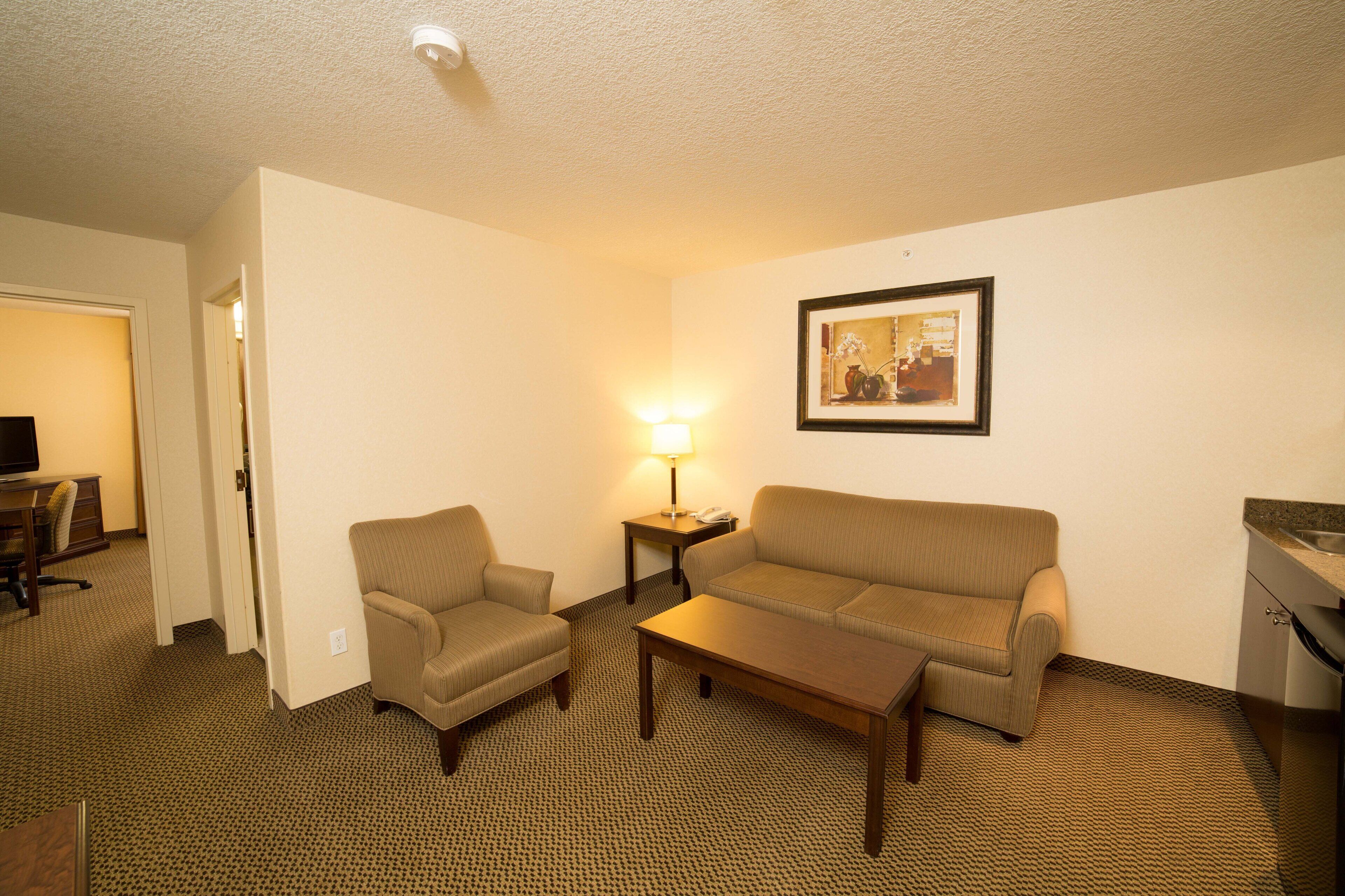 A hotel room with a sofa, armchair, and a small table. There is a lamp on a side table, a framed picture on the wall, and a kitchenette area.