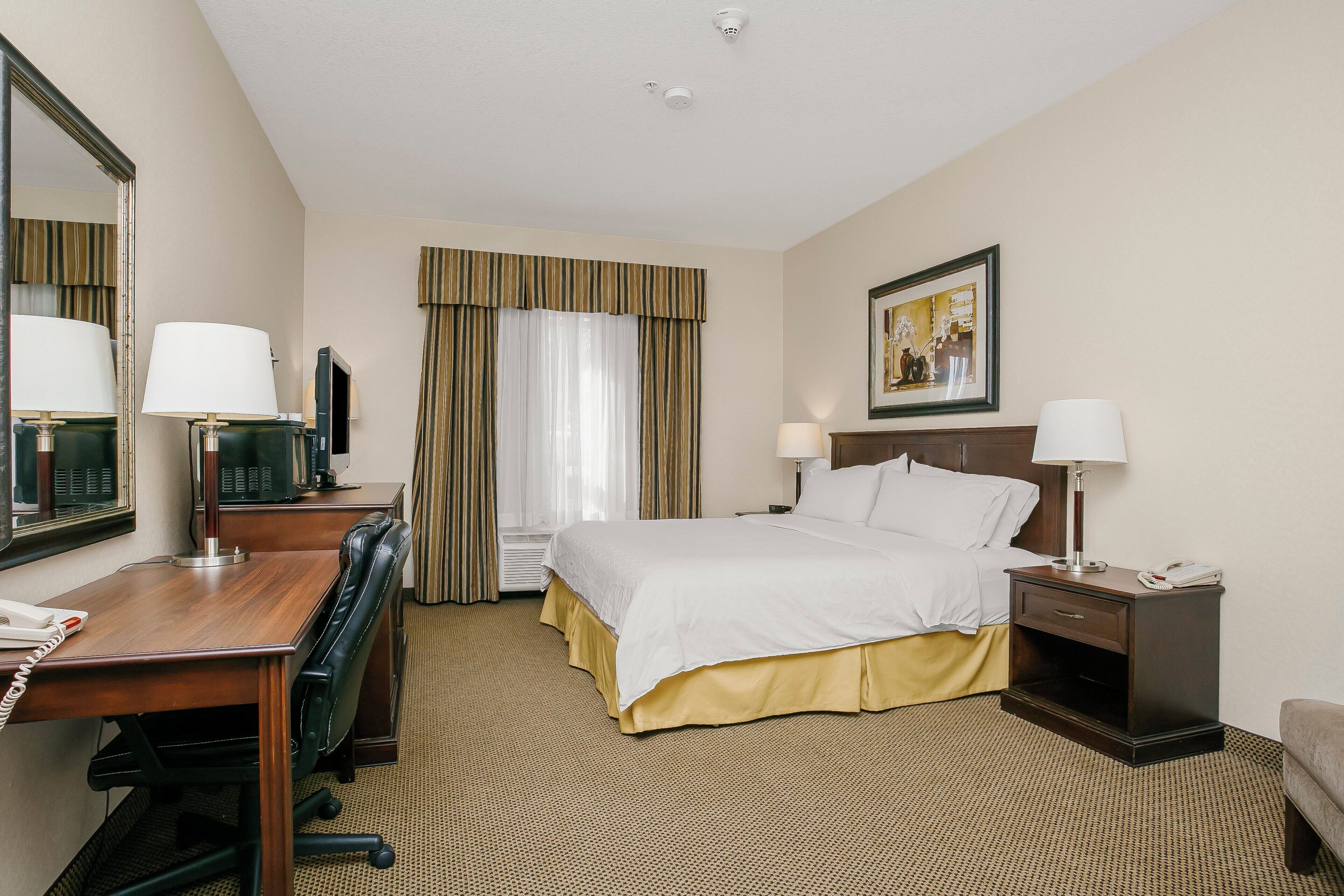 A hotel room with a bed, two bedside tables with lamps, a desk with a telephone, a chair, a large mirror, and a painting on the wall.