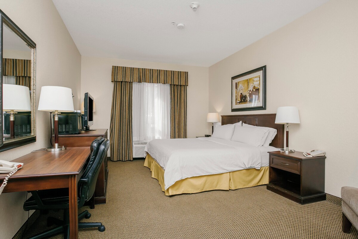 A hotel room with a bed, desk, chair, desk lamp, bedside table, and a painting on the wall.