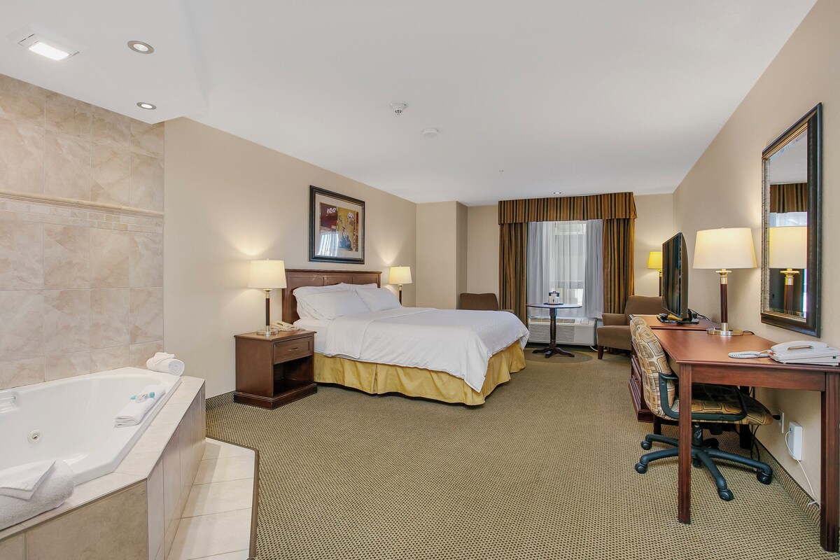 A hotel room with a large bed, a desk, a chair, a desk lamp, a TV, a window with curtains, a bathroom with a bathtub, and a mirror.