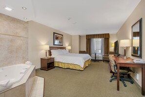 Habitación de hotel con una cama grande, un escritorio, una silla, una lámpara de escritorio, un televisor, una ventana con cortinas, un baño con bañera y un espejo.