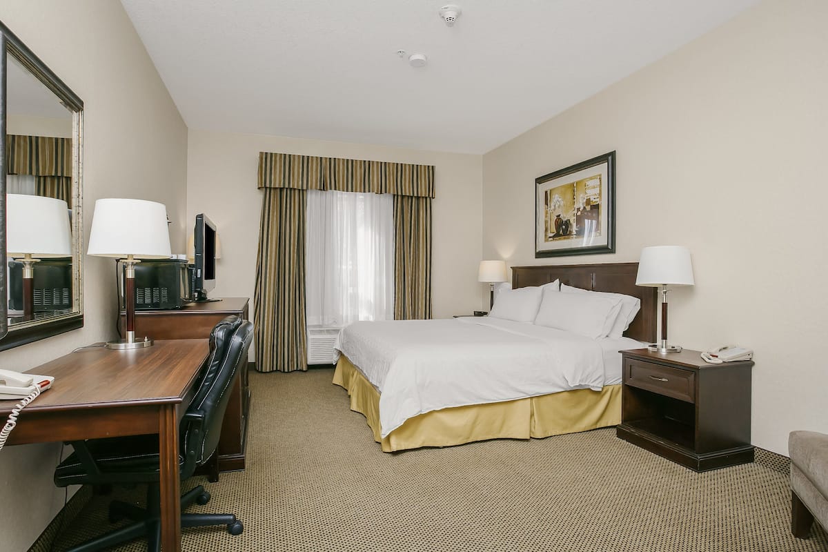 A hotel room with a bed, desk, chair, desk lamp, bedside table, and a painting on the wall.