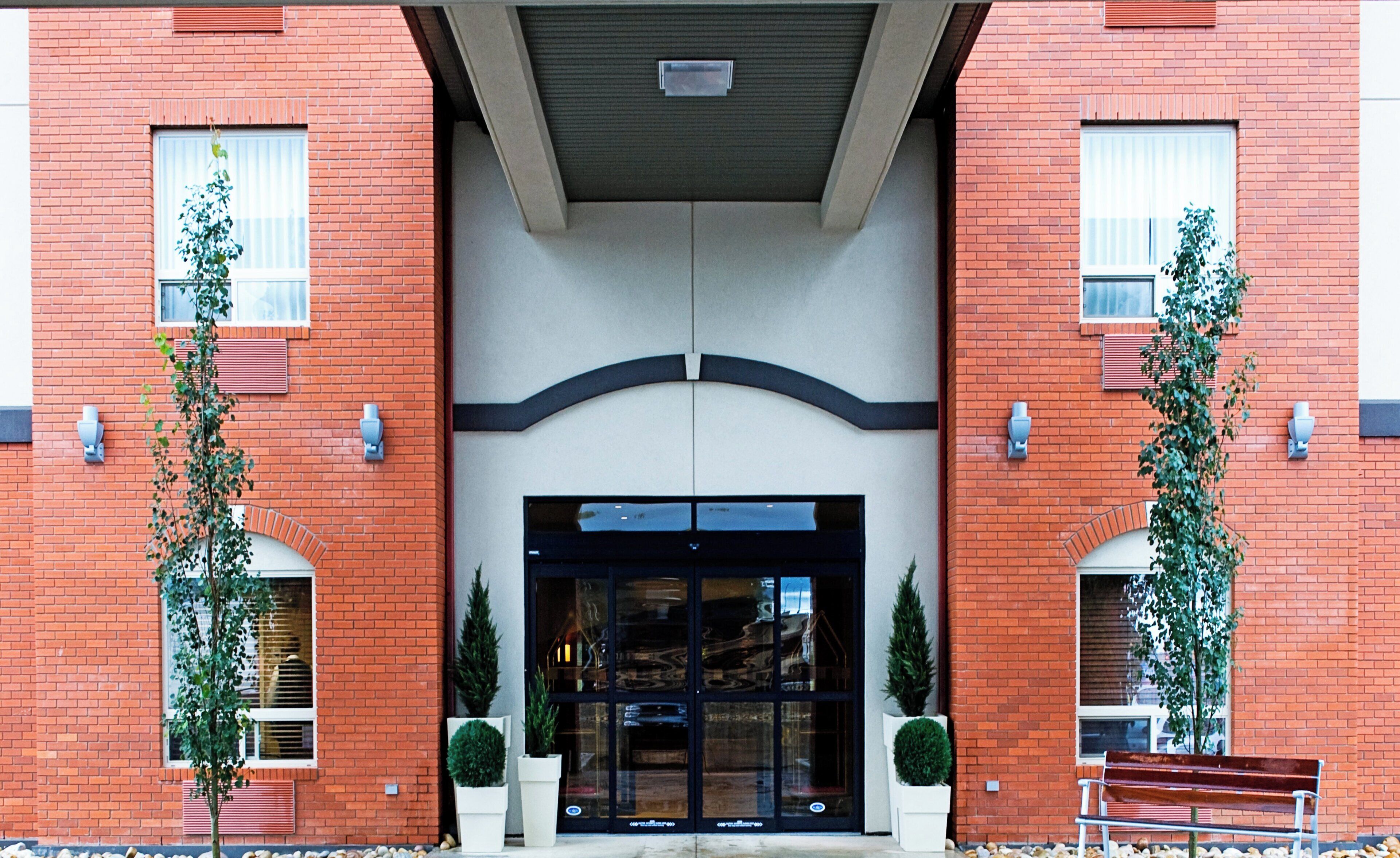 A red brick building with a central entrance, two potted plants on either side, and a bench in front.