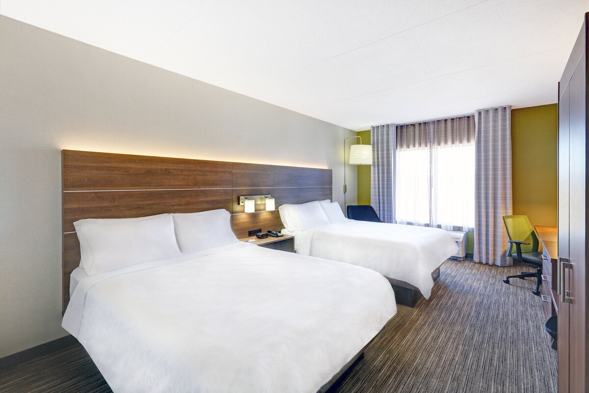 Habitación de hotel con dos camas, un escritorio con silla, una lámpara y una ventana con cortinas.