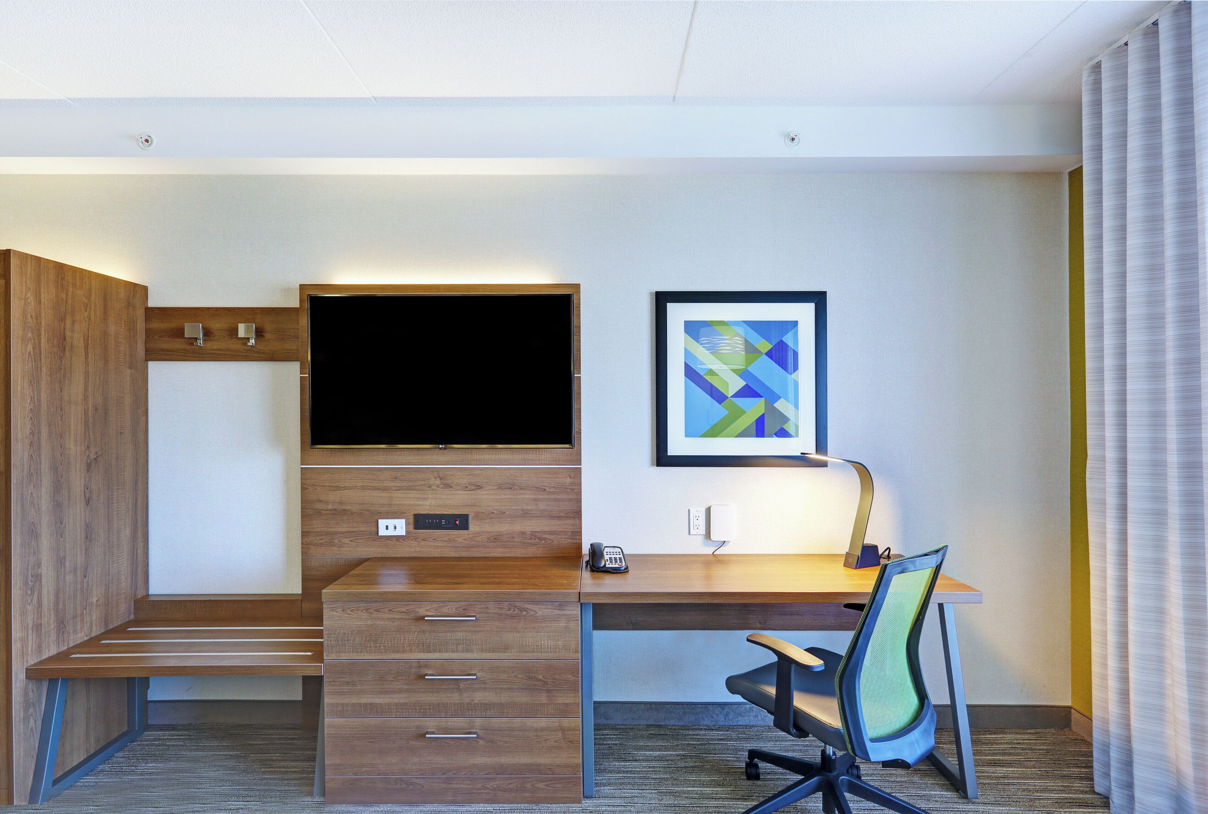 A hotel room with a wooden desk, a flat-screen TV, a framed abstract painting, a telephone, and a chair.