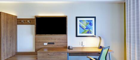 A hotel room with a wooden desk, a flat-screen TV, a framed abstract painting, a telephone, and a chair.