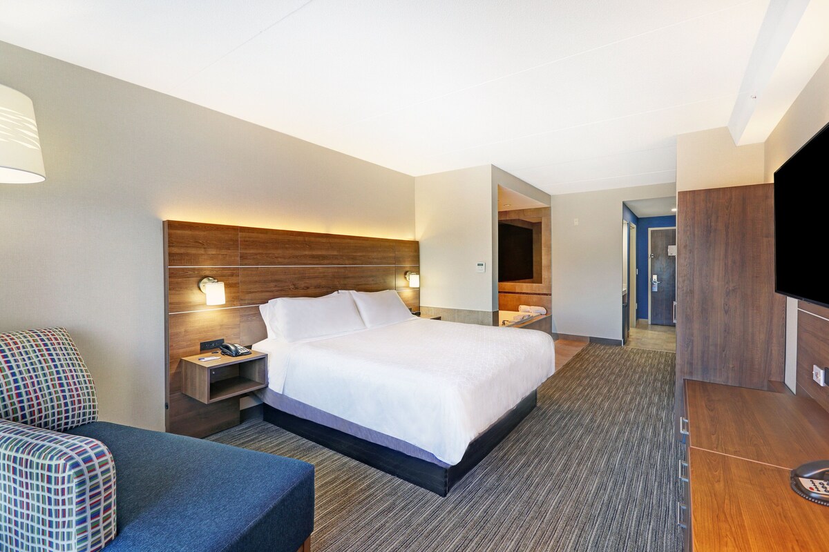 Habitación de hotel con una cama grande, un escritorio con lámpara, televisor y zona de estar con una silla estampada.