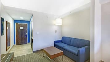A hotel room with a blue sofa, a wooden coffee table, a lamp, and a door leading to a hallway.