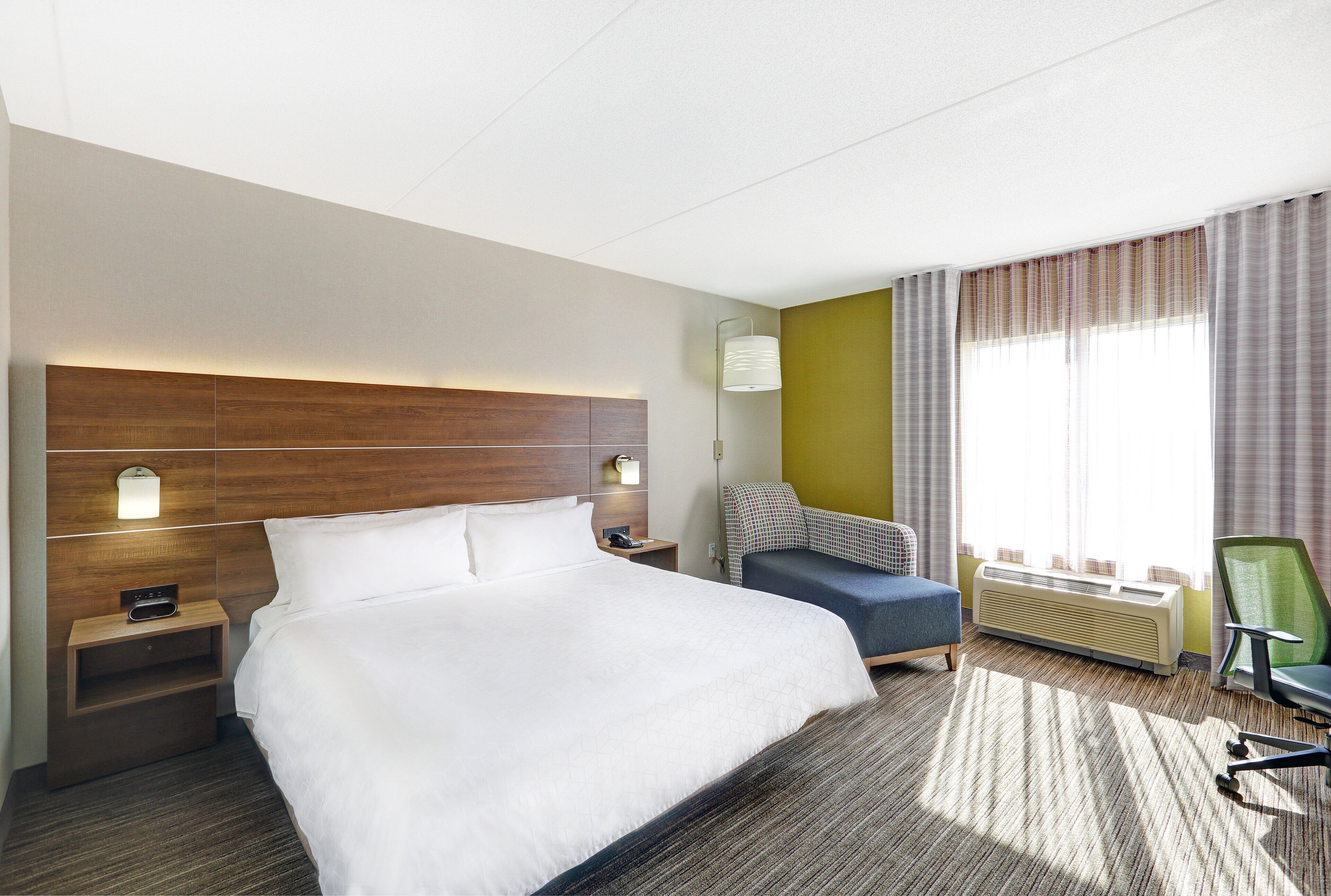 A hotel room with a large bed, a bedside table with a lamp, a chair, a desk, and a window with curtains.