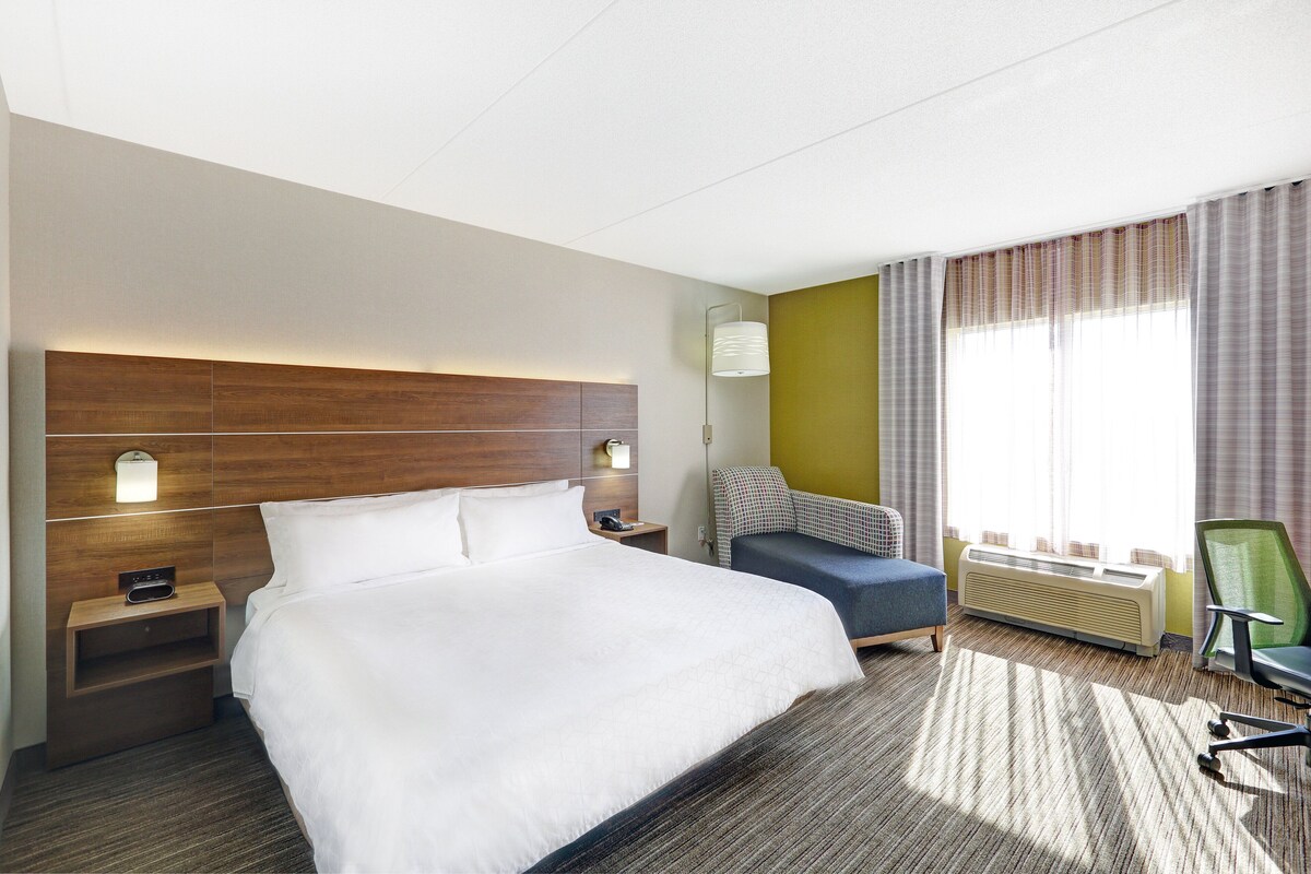 Habitación de hotel con una cama grande, una mesita de noche con lámpara, una silla, un escritorio y una ventana con cortinas.
