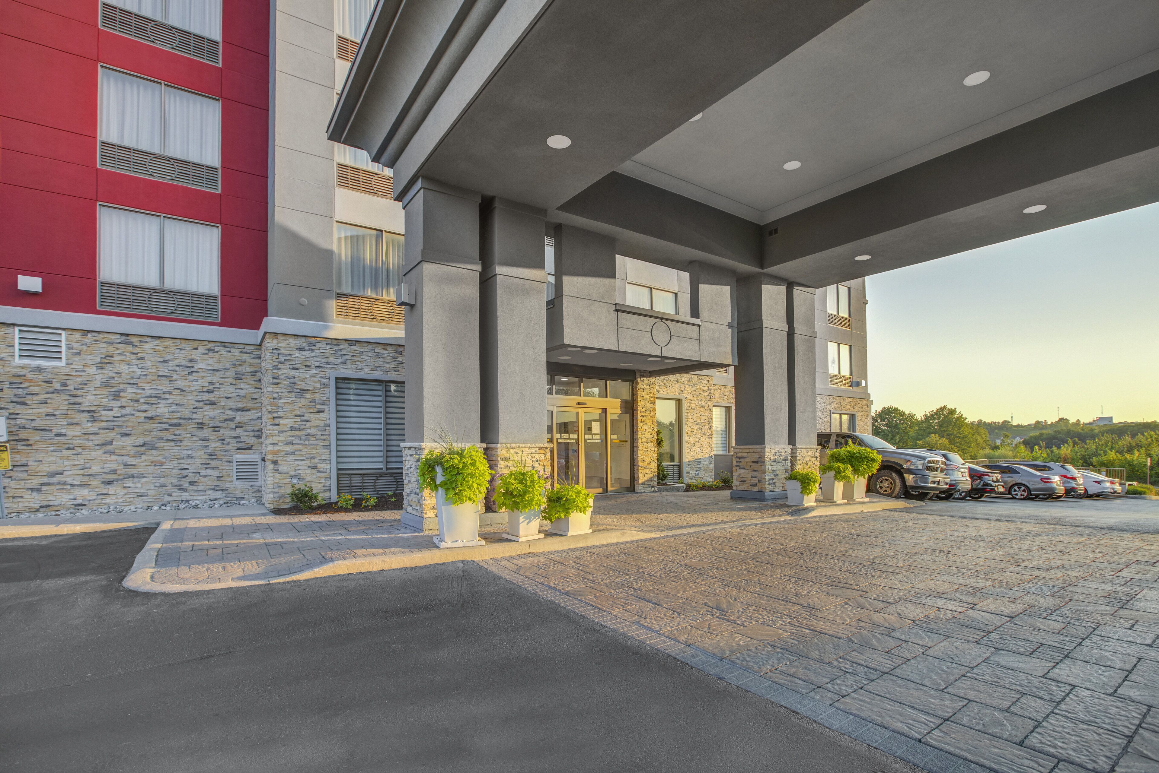 L’entrée d’un hôtel moderne, avec une façade en pierre, un bâtiment rouge sur la gauche et un parking où sont garées plusieurs voitures.