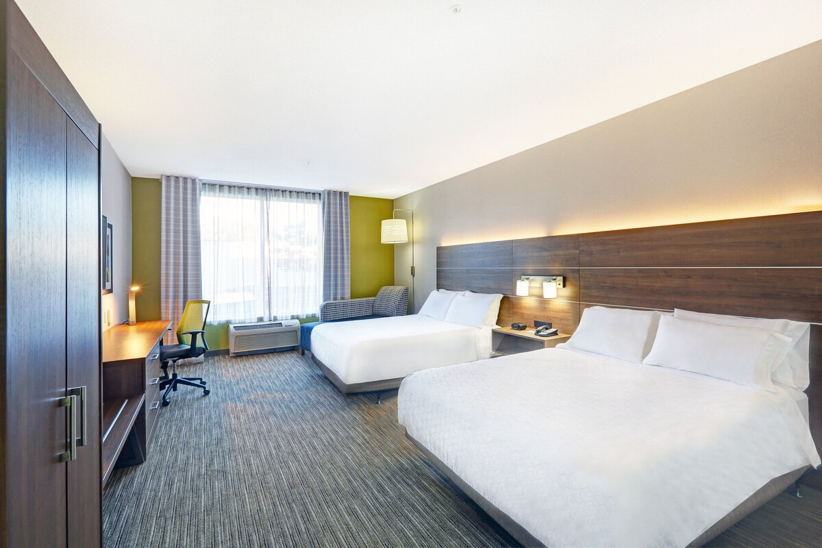 Habitación de hotel con dos camas, un escritorio, una silla, una lámpara y una ventana con cortinas.