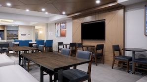 A modern restaurant with wooden tables and blue chairs, a flat-screen TV, and a coffee shop in the background. - Holiday Inn Express Hotel & Suites SWIFT CURRENT by IHG (Swift Current)