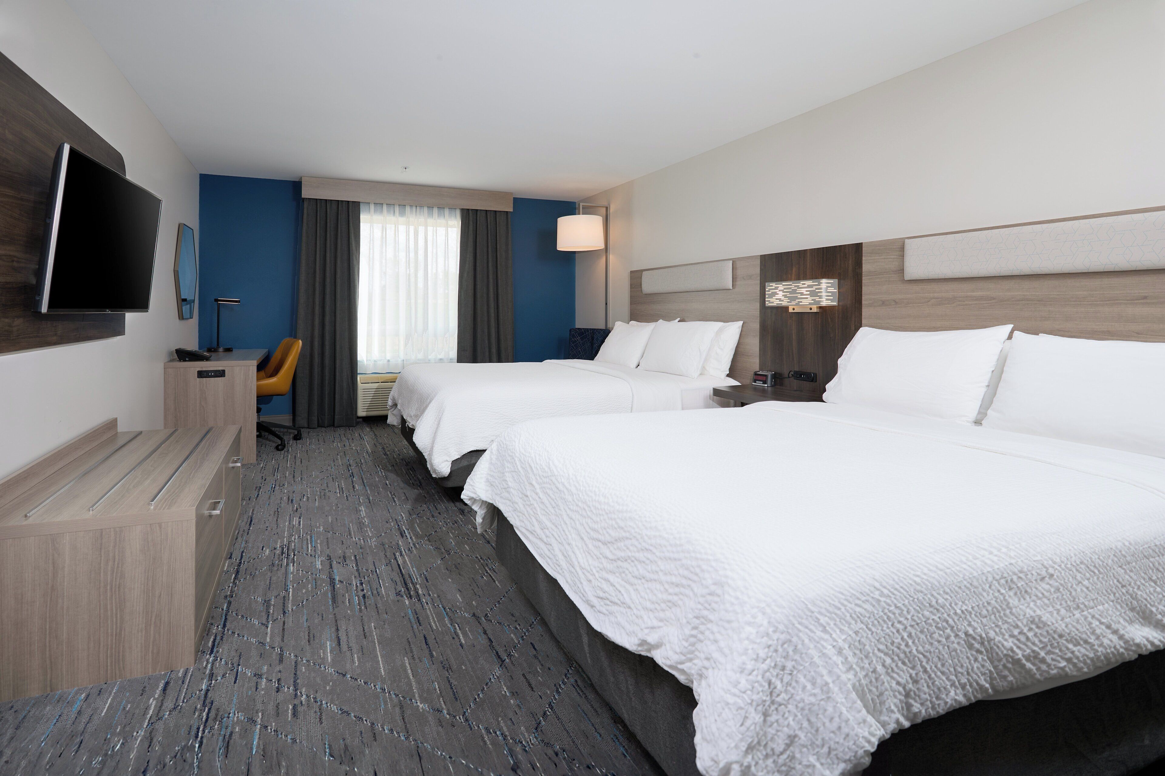 A hotel room with two beds, a desk with a chair, a flat-screen TV, and a large window with curtains.