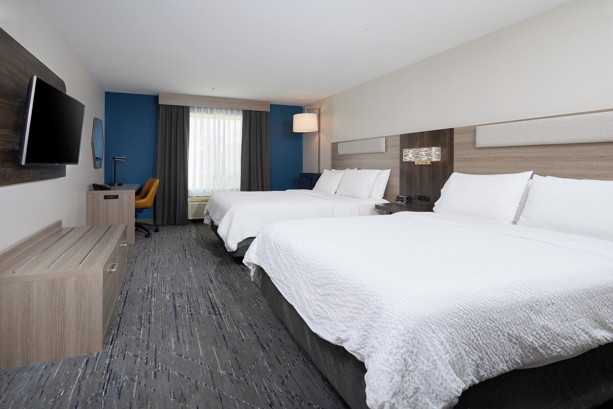 A hotel room with two beds, a desk with a chair, a flat-screen TV, and a large window with curtains.