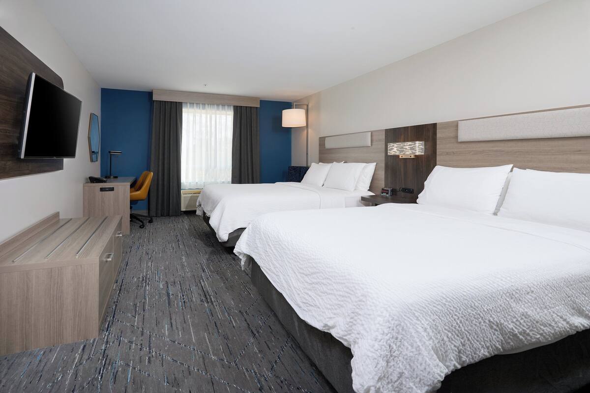 A hotel room with two beds, a desk with a chair, a flat-screen TV, and a large window with curtains.