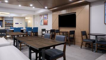 A modern restaurant with wooden tables and blue chairs, a flat-screen TV, and a coffee shop in the background.