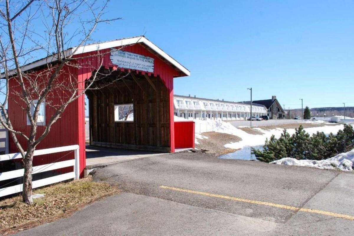 A red covered bridge with a sign, a white building in the background, and a snowy landscape.
