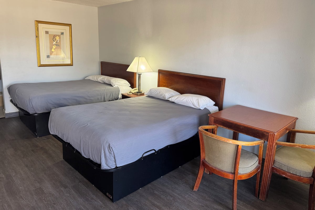 A hotel room with two beds, a desk, a chair, a lamp, and a framed picture on the wall.