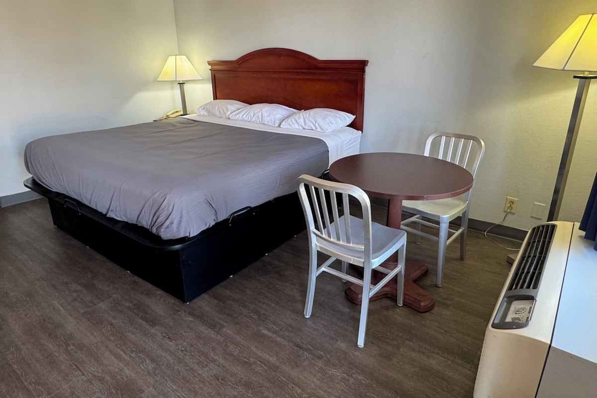 A hotel room with a bed, two chairs, a table, and a heater.