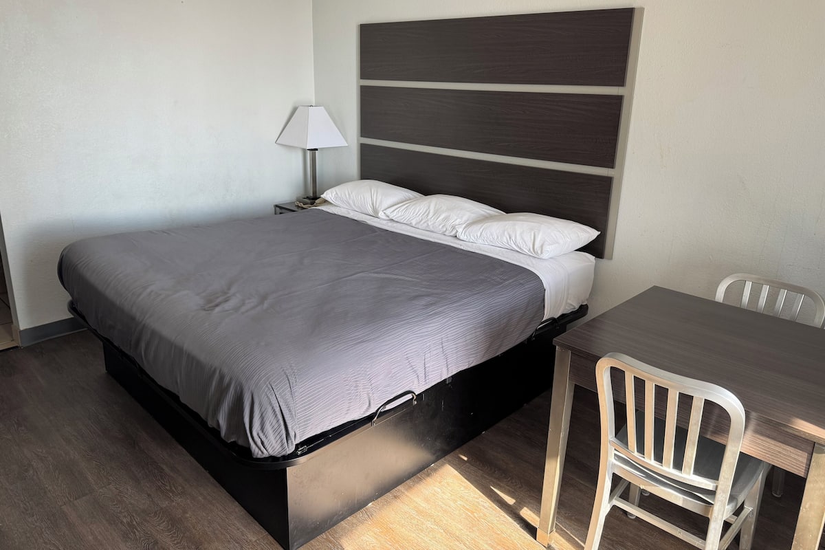 A modern hotel room with a bed, bedside table, lamp, and a small desk with a chair.