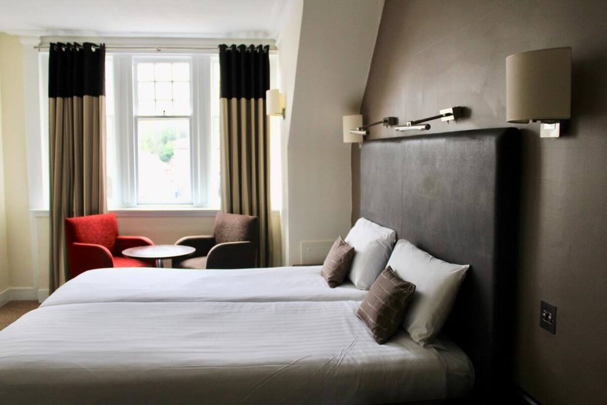 A hotel room with two beds, a red armchair, a small round table, and a window with curtains.