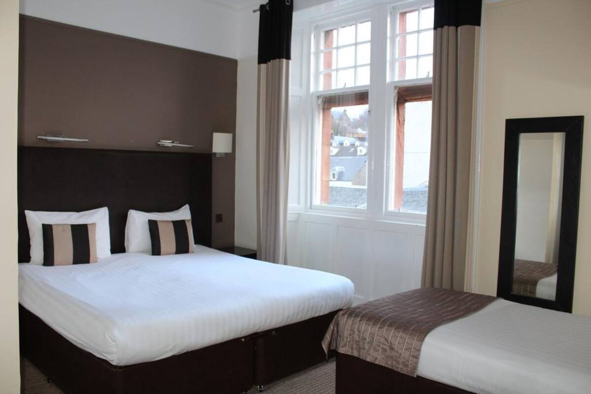 A hotel room with two beds, a large window with curtains, a mirror, and a view of the outside.