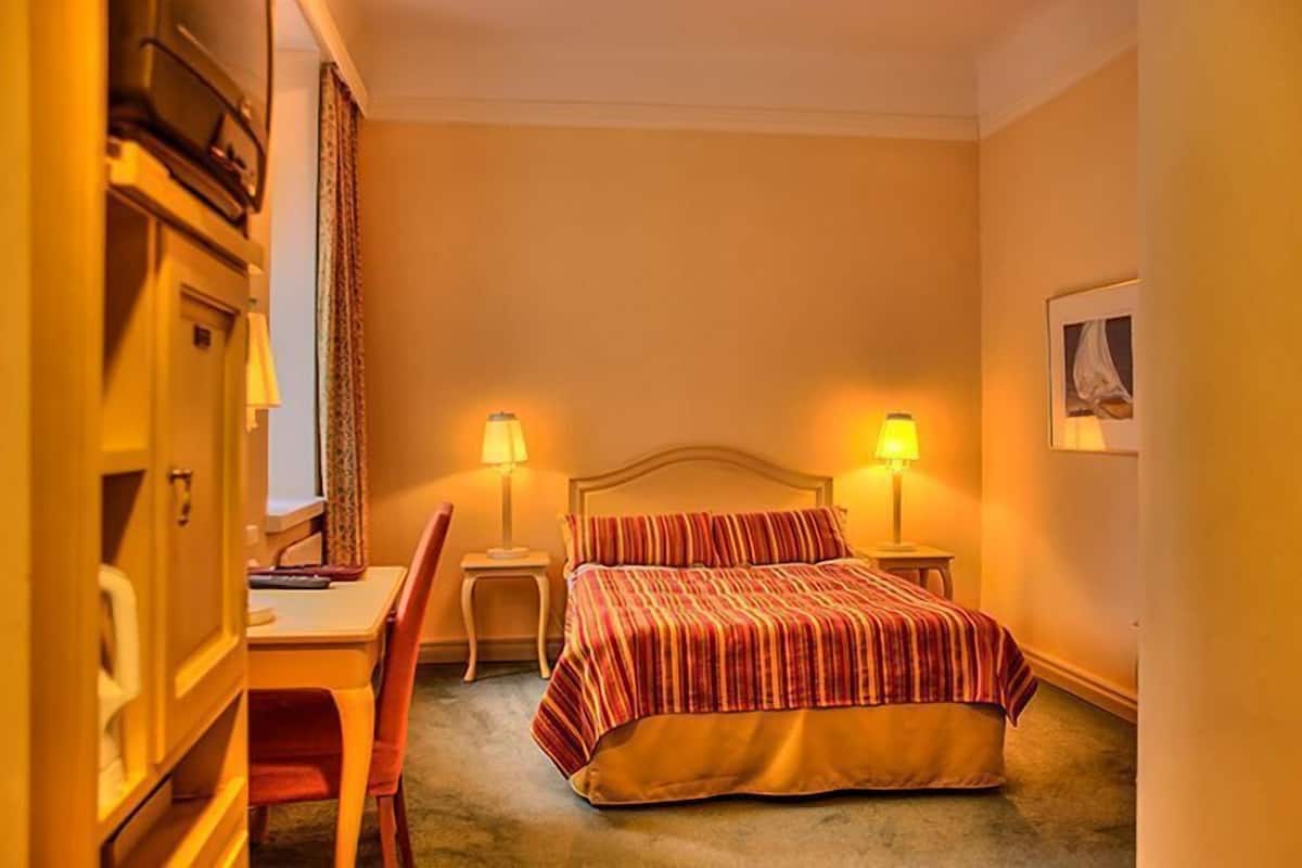 A hotel room with a bed, two bedside lamps, a desk with a chair, and a wardrobe.