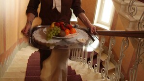 A person carrying a tray with fruits and a cake up a marble staircase with ornate metal railings. - Hotel Astor (Vaasa)